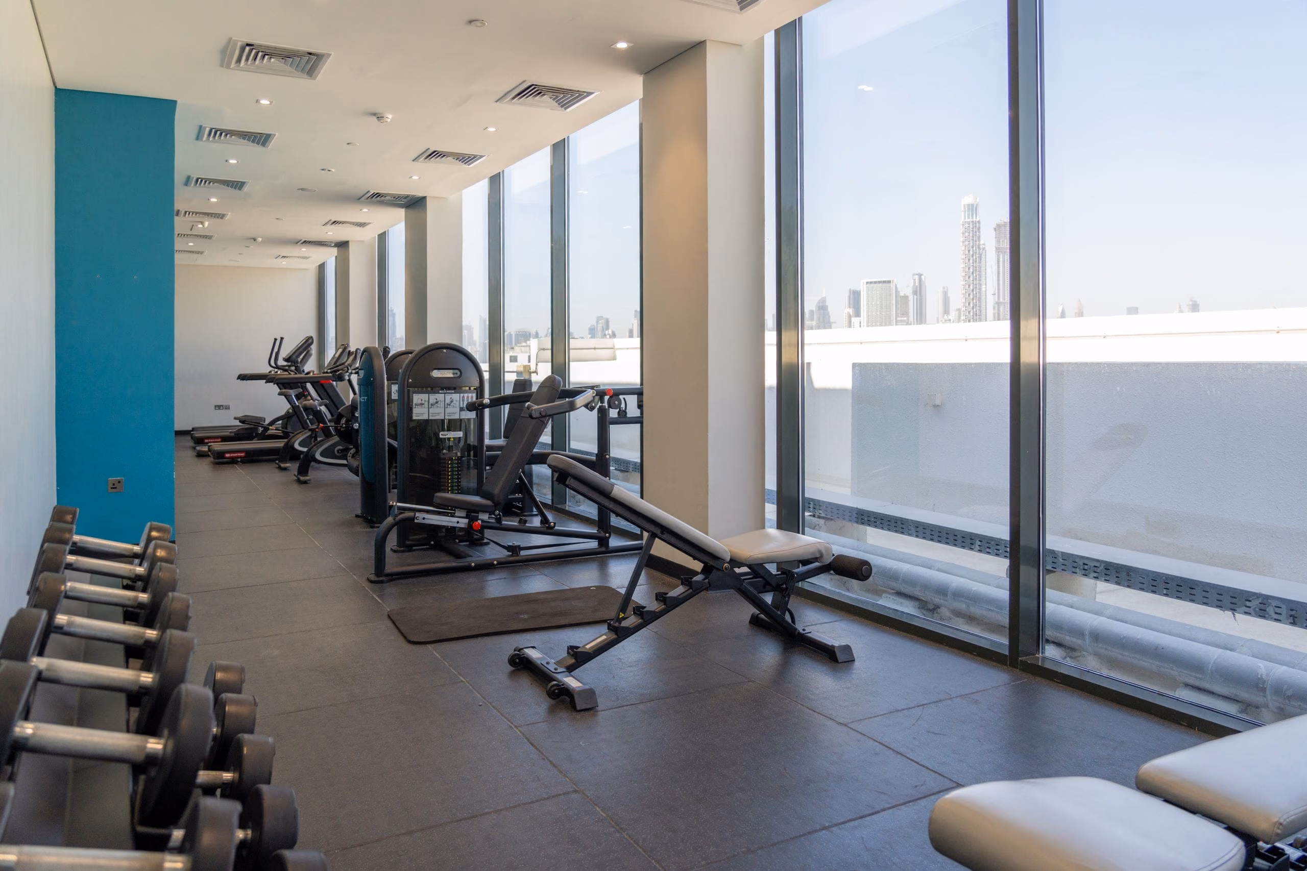 Gym space with floor-to-ceiling glazing, weight bench and machines overlooking a city skyline