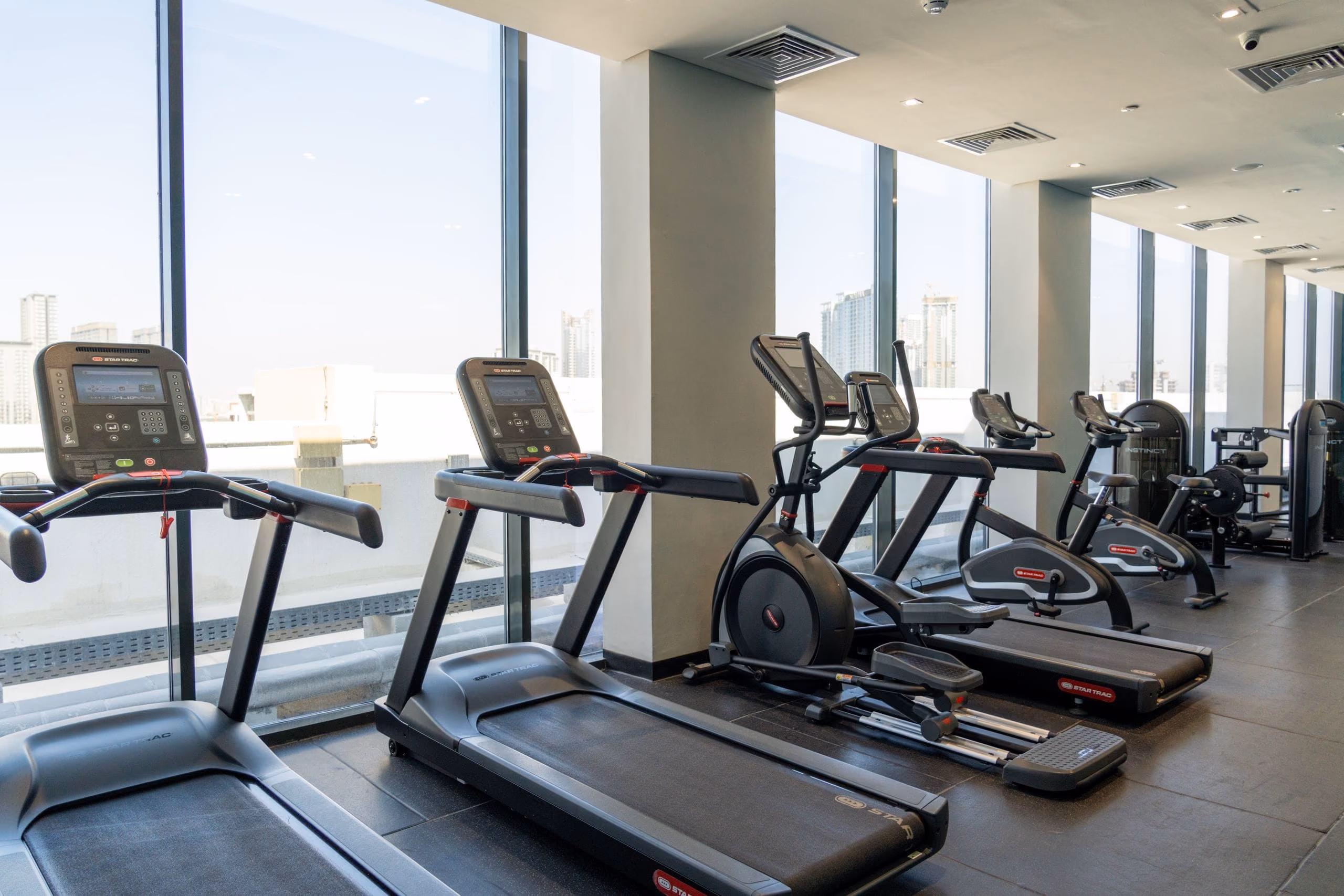 Treadmills and cross trainer lined up along tall glass windows in bright daylight