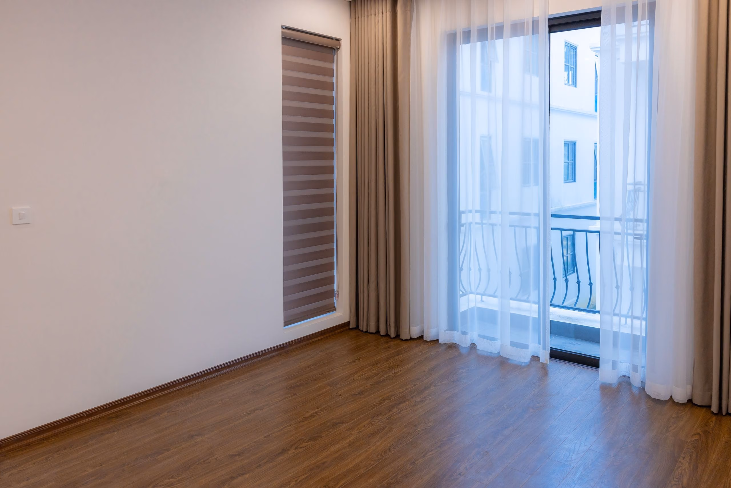 Empty bedroom with new wood plank flooring, sheer curtains and a glass door to a balcony