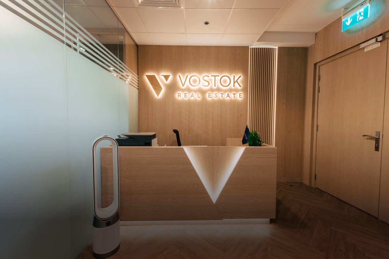 Brend stoli va devordagi yoritilgan logotipga ega Vostok Real Estate ofisi qabulxonasi